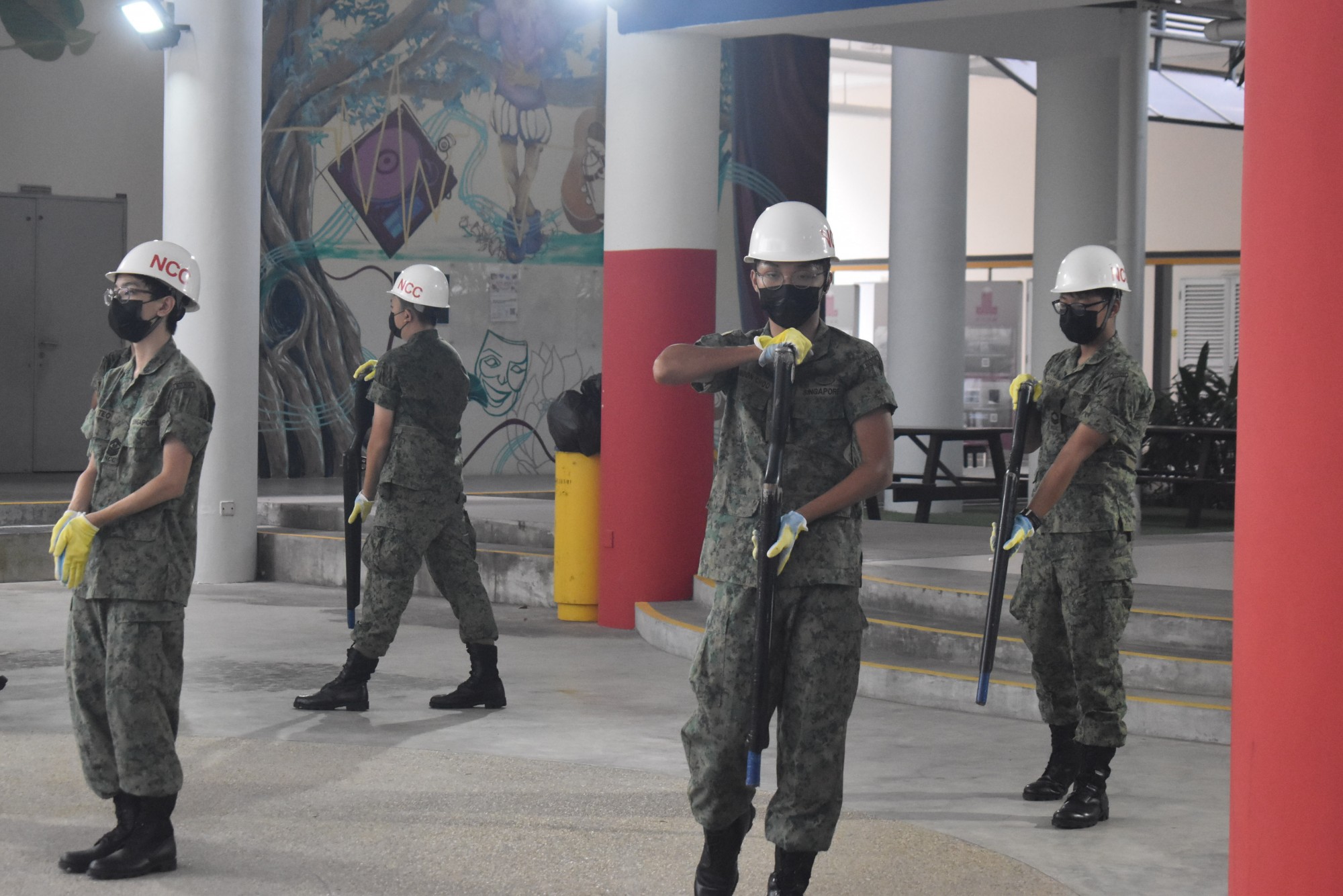 Masked students in military-style uniforms performing a precision drill routine with rifles.